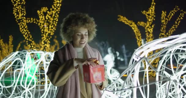年轻的非洲裔美国妇女站在户外的花环和灯光背景上打开礼品盒 新年假期 — 图库视频影像