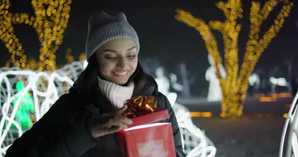 若いインドの女性は ギフトボックスを開き ガーランドに立ってアウトドアのバックグラウンドを明るくします クリスマス休暇 ロイヤリティフリーストック映像