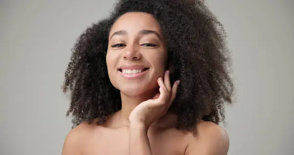 stock image Beauty and healthcare concept - beautiful African American woman with curly afro hairstyle and clean, healthy skin touching face with hand, doing facial massage, posing and looking at camera. High