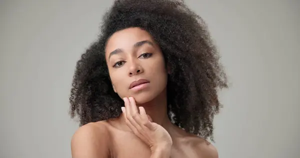 stock image Beauty and healthcare concept - beautiful African American woman with curly afro hairstyle and clean, healthy skin touches her cheek and face with her hand, posing and looking at the camera. High