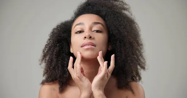 stock image Beauty and healthcare concept - beautiful African American woman with curly afro hairstyle and clean, healthy skin touching face with hand, doing facial massage, posing and looking at camera. High