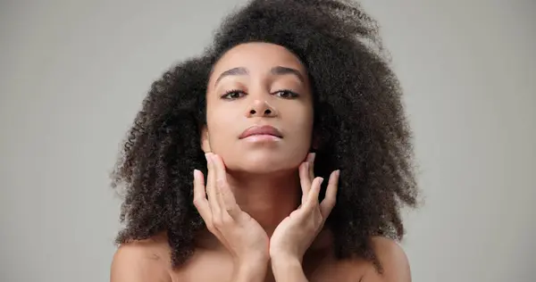 stock image Beauty and healthcare concept - beautiful African American woman with curly afro hairstyle and clean, healthy skin touching face with hand, doing facial massage, posing and looking at camera. High