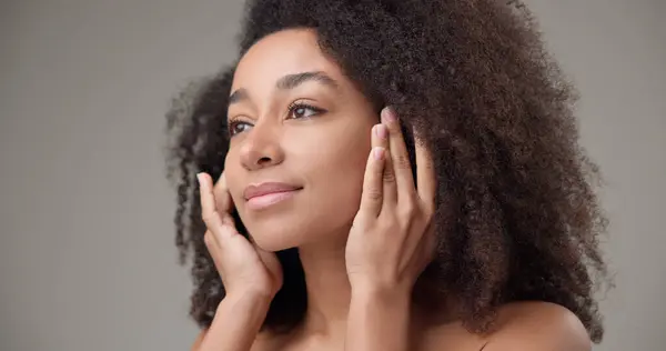 stock image Beauty and healthcare concept - beautiful African American woman with curly afro hairstyle and clean, healthy skin touching face with hand, doing facial massage, posing and looking at camera. High