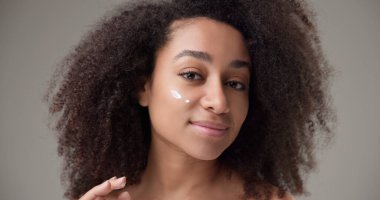 Beauty and healthcare concept - beautiful African American woman with curly afro hairstyle and clean, healthy skin applying moisturizer to her face with a slight smile and positive mood. High quality clipart