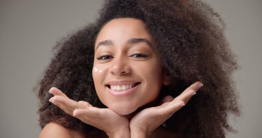 Beauty and healthcare concept - beautiful African American woman with curly afro hairstyle and clean, healthy skin applying moisturizer to her face with a slight smile and positive mood. High quality clipart