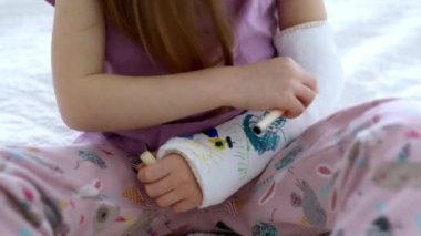 Adorable preschooler girl with a broken arm at home on the bed draws with felt-tip pens on an orthopedic cast. How to have fun with a broken limb. Broken arm in a cast in kids