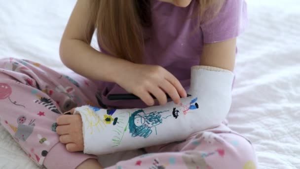 可爱的学龄前女孩 家里的胳膊断了 躺在床上用毛笔在骨科石膏上画画 如何以断肢为乐 孩子们的石膏断了胳膊 — 图库视频影像