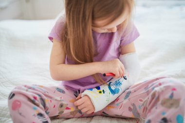 Adorable preschooler girl with a broken arm at home on the bed draws with felt-tip pens on an orthopedic cast. How to have fun with a broken limb. Broken arm in a cast in kids