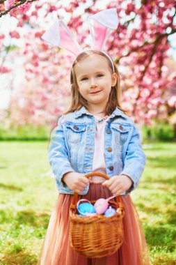 Preschooler girl wearing bunny ears playing egg hunt on Easter. Child gathering colorful eggs in basket. Little kid celebrating Easter outdoors