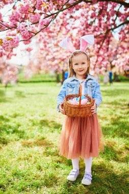 Preschooler girl wearing bunny ears playing egg hunt on Easter. Child gathering colorful eggs in basket. Little kid celebrating Easter outdoors
