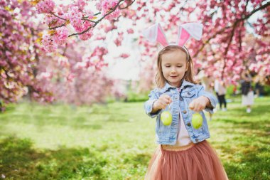 Preschooler girl wearing bunny ears playing egg hunt on Easter. Child gathering colorful eggs in basket. Little kid celebrating Easter outdoors