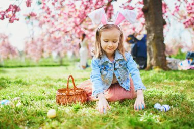 Preschooler girl wearing bunny ears playing egg hunt on Easter. Child gathering colorful eggs in basket. Little kid celebrating Easter outdoors
