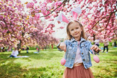 Preschooler girl wearing bunny ears playing egg hunt on Easter. Child gathering colorful eggs in basket. Little kid celebrating Easter outdoors
