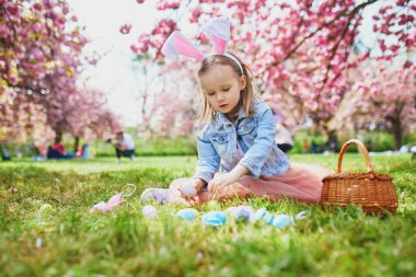 Preschooler girl wearing bunny ears playing egg hunt on Easter. Child gathering colorful eggs in basket. Little kid celebrating Easter outdoors