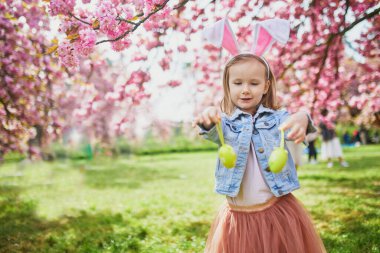 Preschooler girl wearing bunny ears playing egg hunt on Easter. Child gathering colorful eggs in basket. Little kid celebrating Easter outdoors
