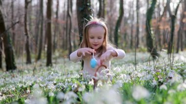 Five year old girl playing egg hunt on Easter. Child sitting on the grass with many snowdrop flowers and gathering colorful eggs in basket. Little kid celebrating Easter outdoors in park or forest