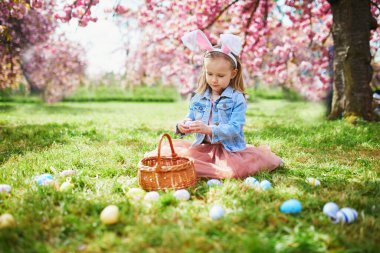 Preschooler girl wearing bunny ears playing egg hunt on Easter. Child gathering colorful eggs in basket. Little kid celebrating Easter outdoors