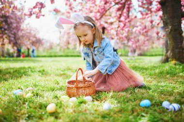Preschooler girl wearing bunny ears playing egg hunt on Easter. Child gathering colorful eggs in basket. Little kid celebrating Easter outdoors