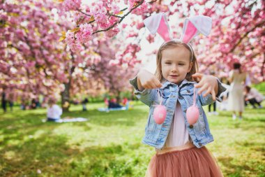 Preschooler girl wearing bunny ears playing egg hunt on Easter. Child gathering colorful eggs in basket. Little kid celebrating Easter outdoors