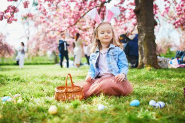 Preschooler girl wearing bunny ears playing egg hunt on Easter. Child gathering colorful eggs in basket. Little kid celebrating Easter outdoors