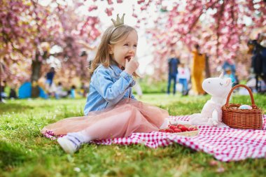 Adorable preschooler girl in tutu skirt and princess crown enjoying picnic with toy unicorn on a nice spring day in cherry blossom garden