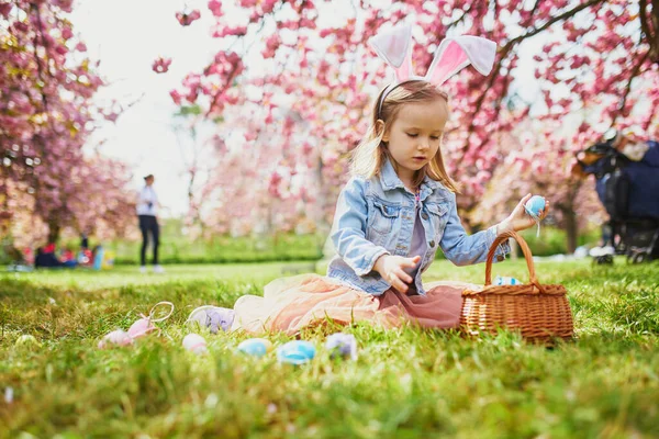 Preschooler girl wearing bunny ears playing egg hunt on Easter. Child gathering colorful eggs in basket. Little kid celebrating Easter outdoors