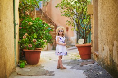 Adorable preschooler girl in white dress and straw hat on a street of Bonnieux village in Provence, France