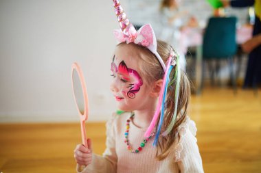 Children face painting. Little preschooler girl in unicorn costume admiring her facepaint on a birthday party. Creative activities for kids clipart