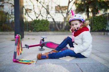 Tek boynuzlu at kasklı anaokulu öğrencisi bir bahar günü parkta scooterını sürerken düştükten sonra yerde oturuyor. Açık hava spor aktiviteleri ve güvenlik kavramı