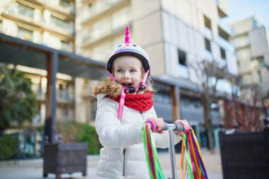 Güneşli bir bahar gününde şehir parkında scooterıyla gezen sevimli anaokulu kızı. Tekboynuz kasklı şirin çocuk scooter kullanıyor. Çocuklar için aktif eğlence ve açık hava sporu