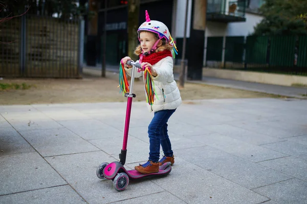 Güneşli bir bahar gününde şehir parkında scooterıyla gezen sevimli anaokulu kızı. Tekboynuz kasklı şirin çocuk scooter kullanıyor. Çocuklar için aktif eğlence ve açık hava sporu