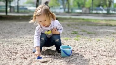 Sevimli küçük kız kova ve kürekle oynuyor, çamur yapıyor ve küçük taşlar topluyor. Çocuklar için açık hava yaratıcı aktiviteler