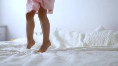 Pembe elbiseli anaokulu kızı eğleniyor ve evdeki yatakta zıplıyor.