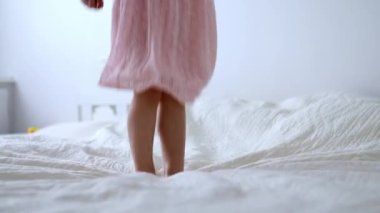 Pembe elbiseli anaokulu kızı eğleniyor ve evdeki yatakta zıplıyor.
