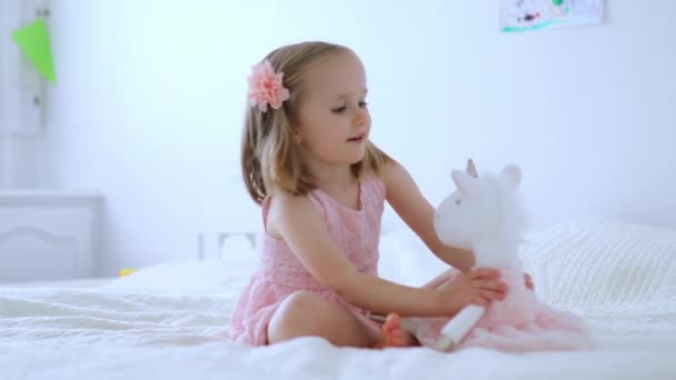 ユニコーンと遊んでピンクのドレスを着た愛らしい女の子 柔らかいおもちゃで楽しんでいる子供 ぬいぐるみで遊ぶ子供 — ストック動画