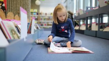 Sevimli anaokulu kızı belediye kütüphanesinde yerde oturmuş kitap okuyor.