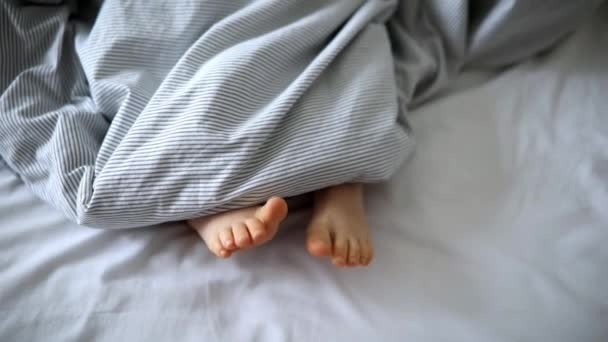 毛布で覆われたベッドの小さな子供の足 快適なベッドで寝ている子供 自宅で早朝に — ストック動画