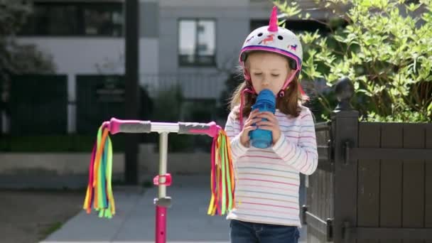 阳光明媚的夏日 一个可爱的学龄前女孩骑着她的摩托车在一个城市公园里 可爱的孩子戴着独角兽头盔喝水 儿童的积极休闲活动和户外运动 — 图库视频影像