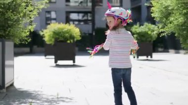 Güneşli bir bahar gününde şehir parkında scooterıyla gezen sevimli anaokulu kızı. Tekboynuz kasklı şirin çocuk scooter kullanıyor. Çocuklar için aktif eğlence ve açık hava sporu
