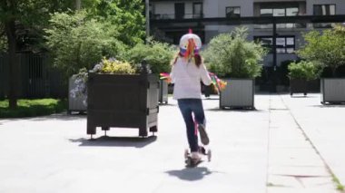 Güneşli bir bahar gününde şehir parkında scooterıyla gezen sevimli anaokulu kızı. Tekboynuz kasklı şirin çocuk scooter kullanıyor. Çocuklar için aktif eğlence ve açık hava sporu