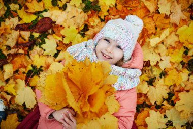 Fransa, Paris 'teki sonbahar parkında düşmüş akçaağaç yapraklarıyla kaplı yerde yatan mutlu küçük kız.