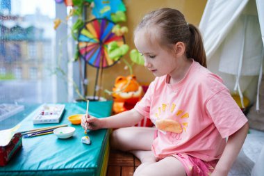 Parlak akrilik boyayla çakıl taşlarını çizen sevimli anaokulu kızı.