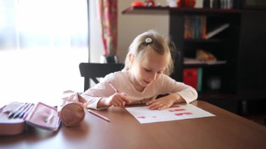 7 yaşında bir kız, evde bir masada oturuyor ve ödevini konsantre bir şekilde yapıyor. Okul malzemeleriyle çevrili, kağıt üzerine kalemle yazıyor.