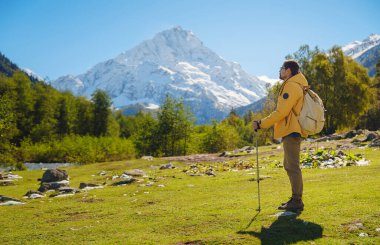 Irkis Vadisi 'nden, Arkhyz' den, Karachay-Cherkessia 'dan, Kuzey Kafkasya' dan. Mavi gökyüzü, bulutlar ve Kafkasya nehri yakınlarındaki güzel ormanla karlı dağ vadisinde yürüyüş yapan bir adam..