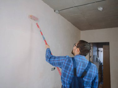 Mavi takım elbiseli genç adam beyaz duvar odası arka planında boya silindiri tutuyor. Yenileme dairesi için aksesuarlar. Ev konseptini onar