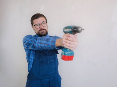Mavi takım elbiseli genç adam apartman dairesinde çalışıyor. Ev yenileme konsepti. Elinde elektrikli matkap tutan tamirci beyaz duvar arkasında izole edilmiş. Enstrümanlar aksesuarlar yenileme dairesi