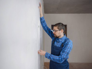 Ciddi bir adamın yer altı ısıtma boruları olan bir odada mezura kullanmasının yan görüntüsü. Tişört tasarımcısı, dairenin tadilatı sırasında ölçü alıyor..