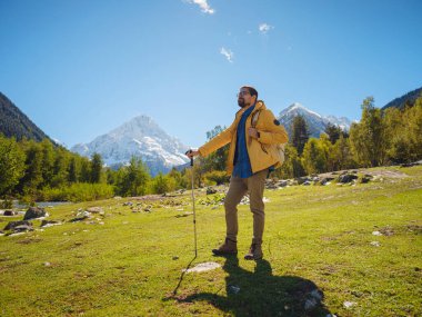 Irkis Vadisi 'nden, Arkhyz' den, Karachay-Cherkessia 'dan, Kuzey Kafkasya' dan. Mavi gökyüzü, bulutlar ve Kafkasya nehri yakınlarındaki güzel ormanla karlı dağ vadisinde yürüyüş yapan bir adam..