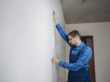 Ciddi bir adamın yer altı ısıtma boruları olan bir odada mezura kullanmasının yan görüntüsü. Tişört tasarımcısı, dairenin tadilatı sırasında ölçü alıyor..