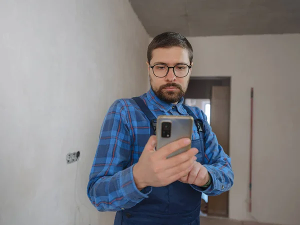 young man in blue work suit doing Repair apartment. Home renovation concept. Guy with smartphone texting and tapping on cellphone, searching for solution on Internet, ordering goods for repairs